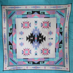 May include: A square, turquoise bandana with a white center featuring a geometric pattern in black, pink, and light blue. The border has a similar design. The bandana is likely made of cotton.