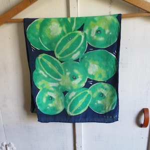 Puede incluir: Un pañuelo azul marino con un patrón repetido de sandías verdes. Las sandías son de varios tamaños y orientaciones, con detalles en blanco. El pañuelo está colgado de una percha de madera.