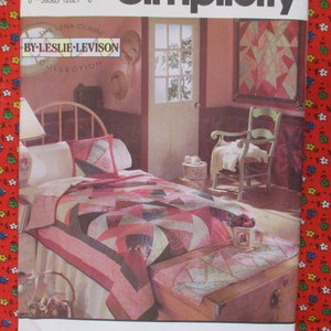 May include: A pink and white quilt with a patchwork design is on a bed in a room with a pink wall. The quilt is made with different fabrics and has a geometric pattern. The bed has a white bedspread and a white pillow with a patchwork design. There is a wooden chair with a white cushion in the corner of the room. The room has a window with a view of trees. The image is from a quilting pattern book.