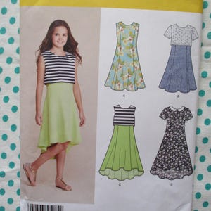 May include: A sewing pattern for girls' dresses. The pattern features a girl wearing a green dress with a striped top. The pattern also shows illustrations of different dress styles, including floral and patterned designs.