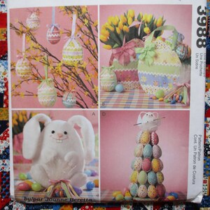 May include: A colorful Easter-themed image featuring decorative eggs, a plush bunny, and a floral arrangement. The image includes hanging eggs, a bunny-shaped egg display, and a tote bag. The background is a patchwork quilt.