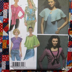 May include: A sewing pattern featuring illustrations of women's tops, including a pink wrap top, a green blouse, and a blue lace cape. The pattern includes a barcode and a colorful patchwork fabric background.