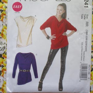 May include: A sewing pattern for a women's top with a cowl neck. The pattern includes three different views: a cream top, a red top, and a purple top. The pattern is labeled "Easy" and includes the size range 18W-20W-22W-24W.
