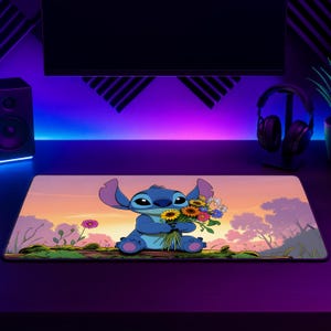 May include: A rectangular desk mat featuring a blue alien character sitting on a log and holding a colourful bouquet of yellow, orange, and white flowers against a sunset background with silhouettes of trees and a pinkish-orange sky.