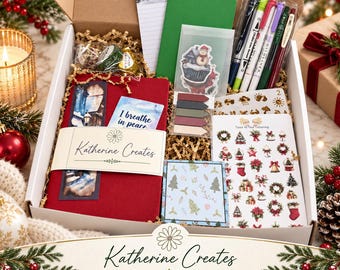 May include: A holiday stationery gift box containing a red journal with a Katherine Creates belly band, a green notebook, a set of colorful markers, a snowman sticker, sticky tabs, a sheet of Christmas-themed stickers, and a notepad with winter art.