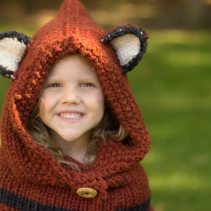 May include: A knitted fox hood in shades of rust and brown, featuring black stripes and a wooden button closure. The hood has fox ear details with white and black accents. The child is smiling, wearing the hood outdoors.