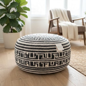 May include: A round, crocheted pouf featuring a black and white geometric pattern. The textured, cylindrical ottoman has a flat top and a small white fabric tag on the side with the text CRCHTHS STUDIO printed in black, centered on the side panel.