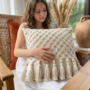 May include: A square throw pillow featuring a cream-colored fabric base with an intricate macrame overlay in a diamond lattice pattern. The bottom edge is adorned with a row of light wooden beads and matching cream-colored fabric tassels.