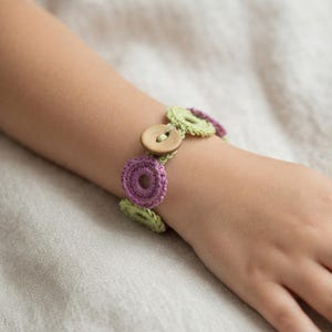 May include: A bracelet featuring alternating light green and purple crocheted circular motifs connected together. The bracelet is secured with a single round, light brown wooden button that acts as a functional closure for the piece.