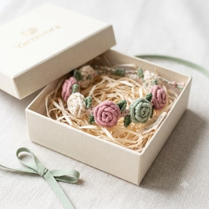 May include: A crochet bracelet featuring small pink, cream, and sage green yarn roses with green leaves, nestled in a beige cardboard gift box filled with wood shavings. The box lid is labeled with the text Yarnivora and a sage green ribbon.