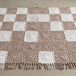 Può includere: Una coperta quadrata all'uncinetto con un motivo a scacchiera di quadrati alternati in filato bianco e beige. La coperta è composta da singoli quadrati uniti tra loro ed è rifinita con frange lungo tutti e quattro i bordi.