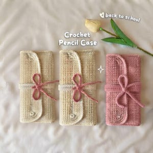 May include: Three rectangular crochet pencil cases, two in cream and one in dusty pink, each featuring a wrap-around closure secured with a matching crocheted tie. The text "Crochet Pencil Case" and "back to school" appears above the items.
