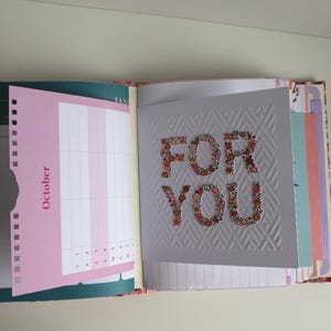 Può includere: Un'agenda aperta che mostra una pagina rosa etichettata October con una griglia e un inserto in cartoncino bianco con un motivo geometrico in rilievo e la scritta FOR YOU in stampa floreale. L'agenda include vari divisori e pagine colorate.