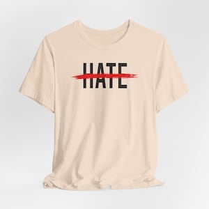 May include: A light beige short-sleeved t-shirt featuring the word HATE printed in bold black capital letters across the chest, with a horizontal red brushstroke graphic drawn directly through the center of the text.