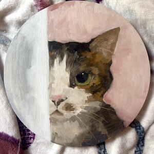 Könnte beinhalten: Ein kreisförmiges Ölgemälde auf Leinwand, das ein Nahporträt einer Katze mit braunem, weißem und schwarzem Fell zeigt. Die Katze hat große grüne Augen und eine rosa Nase. Der Hintergrund ist in einen vertikalen weißen Streifen und einen rosa Bereich unterteilt.