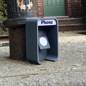 Puede incluir: Un soporte de carga gris mate para reloj inteligente, de forma rectangular, con un diseño de cabina telefónica, la palabra Phone impresa en azul sobre fondo blanco en la parte superior y un disco de carga blanco circular en el centro.
