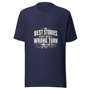 May include: A navy blue short-sleeve t-shirt featuring a graphic print in white. The text reads THE BEST STORIES START WITH A WRONG TURN above an illustration of a winding road through a forest with a silhouette of a creature walking.