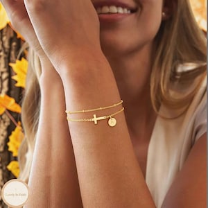 May include: A gold-toned double-strand bracelet featuring a delicate chain with small beads and a second chain with a horizontal cross charm and a small circular pendant engraved with the letter A. The jewelry has a polished metallic finish.
