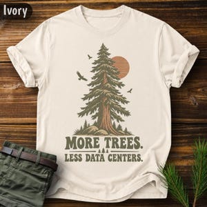 Puede incluir: Una camiseta de manga corta color marfil con un gráfico de un gran pino, un sol y pájaros. Debajo del árbol, el texto dice: MORE TREES. LESS DATA CENTERS. El diseño utiliza tonos tierra de verde y marrón para representar la escena forestal.