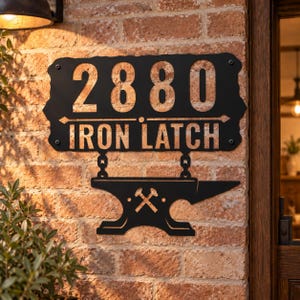 Könnte beinhalten: Ein schwarzes Metall-Wandschild mit der Nummer 2880 und dem Text IRON LATCH. Das Schild enthält eine dekorative Ambossform, die an zwei kleinen Ketten hängt, mit einem Symbol aus gekreuzten Hämmern in der Mitte des Ambosskörpers.