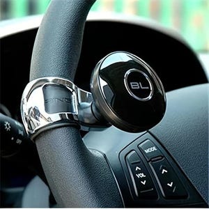 May include: A chrome-finished steering wheel spinner knob featuring a circular black control head with the letters BL printed in white. The device is securely clamped onto the textured black rim of a vehicle steering wheel with integrated controls.