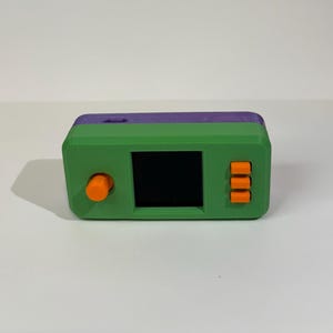 May include: A rectangular handheld electronic device with a green front casing and a purple top section. It features a central black square screen, a single round orange knob on the left side, and three vertical orange rectangular buttons on the right.