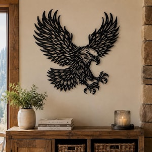 May include: A black metal wall art piece shaped like a bald eagle in flight with wings spread wide and talons extended forward. The eagle features intricate cut-out details throughout its feathers and body, creating a silhouette effect against the wall.