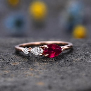 Pode incluir: Um anel de ouro rosa com um rubi vermelho em forma de pera, um diamante branco em forma de pera e um pequeno diamante branco redondo dispostos em fila. O aro é fino e polido, exibindo uma disposição delicada de três pedras preciosas.
