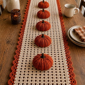 May include: A rectangular crochet table runner featuring a cream-colored grid pattern with a scalloped orange border. Five small, ribbed, orange crocheted pumpkins with dark green stems are arranged in a single line down the center of the runner.