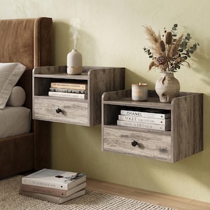 May include: Two wall-mounted floating nightstands with a gray wood grain finish, each featuring an open shelf and a single drawer with a small black knob. The shelves hold books, a small white diffuser, a candle, and a decorative vase with dried plants.