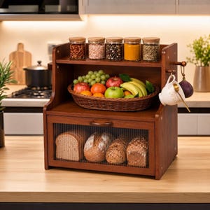 May include: A two-tier wooden bread box with a fluted glass door containing four loaves of bread. The top shelf holds five glass spice jars, a woven basket filled with assorted fresh fruit, and a white teacup hanging from a side hook.