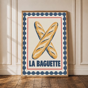 May include: A rectangular wall art print in a wooden frame featuring two crossed golden-brown baguettes illustrated in a painterly style. The design includes a dark blue scalloped border, a thin red inner frame, and the text LA BAGUETTE in blue.