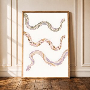 Könnte beinhalten: Ein gerahmter Kunstdruck mit drei horizontal angeordneten Schlangen in Aquarelloptik. Die obere Schlange ist grün und grau, die mittlere ist beige mit braunen Punkten und die untere ist violett und beige. Jede Schlange hat einen gewellten Körper.