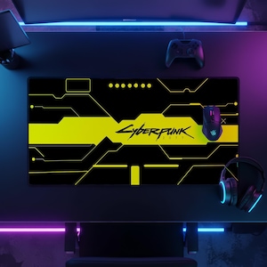 May include: A rectangular desk mat featuring a black and yellow geometric circuit board pattern with the Cyberpunk 2077 logo printed in the centre. The mat is positioned on a dark surface alongside a black computer mouse, a game controller, and headphones.