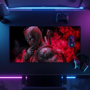 Può includere: Un tappetino da scrivania rettangolare per gaming con un'illustrazione grafica del personaggio Deadpool in costume rosso e nero, su uno sfondo rosso fumoso e vibrante. Il tappetino è posizionato su una scrivania scura con controller e mouse.