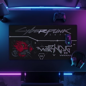 Puede incluir: Una alfombrilla de escritorio rectangular negra con el logotipo de Cyberpunk 2077 en texto plateado en la parte superior. El diseño incluye un gráfico rojo y negro de una máscara oni estilizada, la palabra WARNING en letras blancas y símbolos.