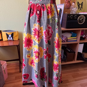 May include: A long, high-waisted skirt featuring a black and white gingham check pattern overlaid with a vibrant floral print. The flowers are rendered in shades of bright red, pink, and yellow. The skirt has an elasticized waistband.
