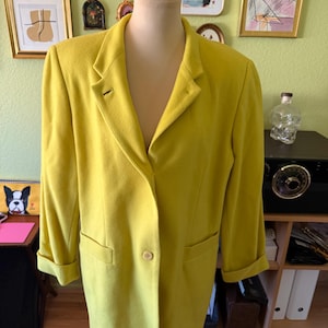 May include: A chartreuse yellow blazer featuring a notched lapel, a single front button closure, and long sleeves with folded cuffs. The garment includes two horizontal welt pockets on the lower front section and is displayed on a mannequin.