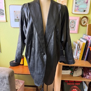 May include: A black leather blazer featuring a single-button closure, wide lapels, and structured shoulders. The jacket has long sleeves with a slightly gathered detail at the shoulder seam and a smooth, matte finish throughout the leather material.