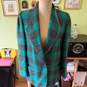 May include: A long-sleeved blazer featuring a bold green and dark gray plaid pattern. The garment has a notched lapel with dark gray trim, slightly puffed shoulders, and a single front button closure with two horizontal slit pockets at the waist.
