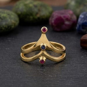 Könnte beinhalten: Ein goldener Ring mit V-förmigem Design und zwei Bändern, die an einem zentralen Punkt zusammenlaufen. Der Ring ist mit drei kleinen, runden, facettierten Edelsteinen besetzt: einem roten Rubin oben, einem blauen Saphir in der Mitte und einem roten Rubin unten.