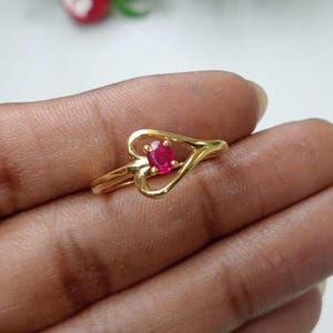 Könnte beinhalten: Ein goldener Ring mit einem herzförmigen Banddesign und einem kleinen, oval geschliffenen, leuchtend roten Edelstein, der sicher in der Mitte gefasst ist. Das polierte goldene Metallband biegt sich um den zentralen roten Stein.