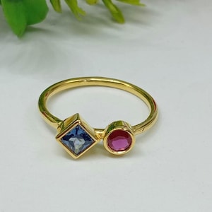 Könnte beinhalten: Ein goldener Ring mit zwei verschiedenen Edelsteinen: einem hellblauen Stein im Quadratschliff und einem leuchtend pinken Stein im Rundschliff. Die Ringschiene ist dünn und poliert und unterstreicht den Kontrast der beiden farbigen Steine.