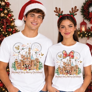 May include: Two white t-shirts featuring a graphic print of a gingerbread castle, Mickey and Minnie Mouse, and the text "Mickey's Very Merry Christmas". The design includes festive holiday elements, golden swirls, and a red bow on the right shirt.