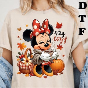 May include: A beige t-shirt featuring a graphic of Minnie Mouse wearing a red and white polka dot bow, holding a steaming mug of coffee. The design includes a wicker basket with flowers and berries, an orange pumpkin, and autumn leaves with the text stay cozy.