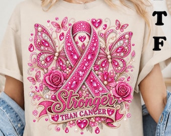 May include: A beige t-shirt featuring a graphic print of a pink ribbon, butterflies, and roses adorned with sparkling pink gems. The design includes a banner with the text Stronger THAN CANCER in pink lettering. The letters DTF appear in black.