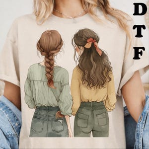 May include: A beige t-shirt featuring a graphic illustration of two individuals seen from behind. One has a long braided hairstyle and wears a sage green top with dark green jeans. The other has wavy brown hair with a red bow and a yellow top.