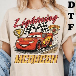 May include: A beige t-shirt featuring a graphic of a red race car with the number 95, a gold trophy, and checkered flags. The text reads Lightning McQueen and Speedway Club 95 Est. 1970. The design includes yellow flame accents and a road background.