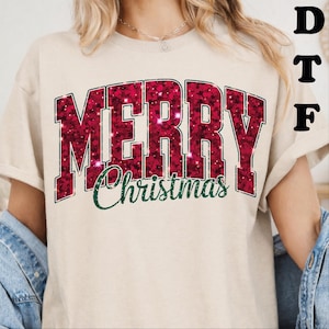 May include: A beige short-sleeved t-shirt featuring the word MERRY in large, block-style letters with a dark red, glittery, textured pattern. Below, the word Christmas is written in a dark green, cursive script font. The letters DTF appear in black.