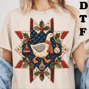 May include: A beige t-shirt featuring a central graphic of a white goose wearing a red and white plaid scarf. The goose is centered within a quilted star pattern in navy blue, red, and cream, decorated with holly berries and Christmas ornaments.
